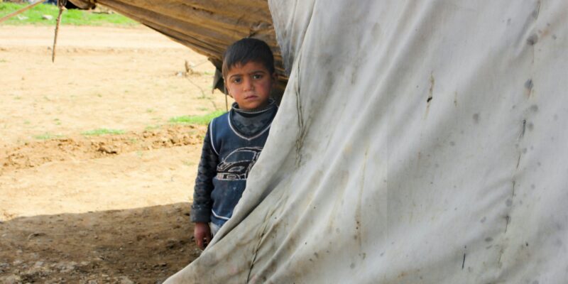 A young boy standing in front of a tent evokes the stark realities of displacement, a poignant connection to Myanmar’s ongoing humanitarian crisis, by Salah Darwish via Unsplash, July 23, 2024, https://unsplash.com/photos/a-young-boy-standing-in-front-of-a-tent-Wti2okH2uvA